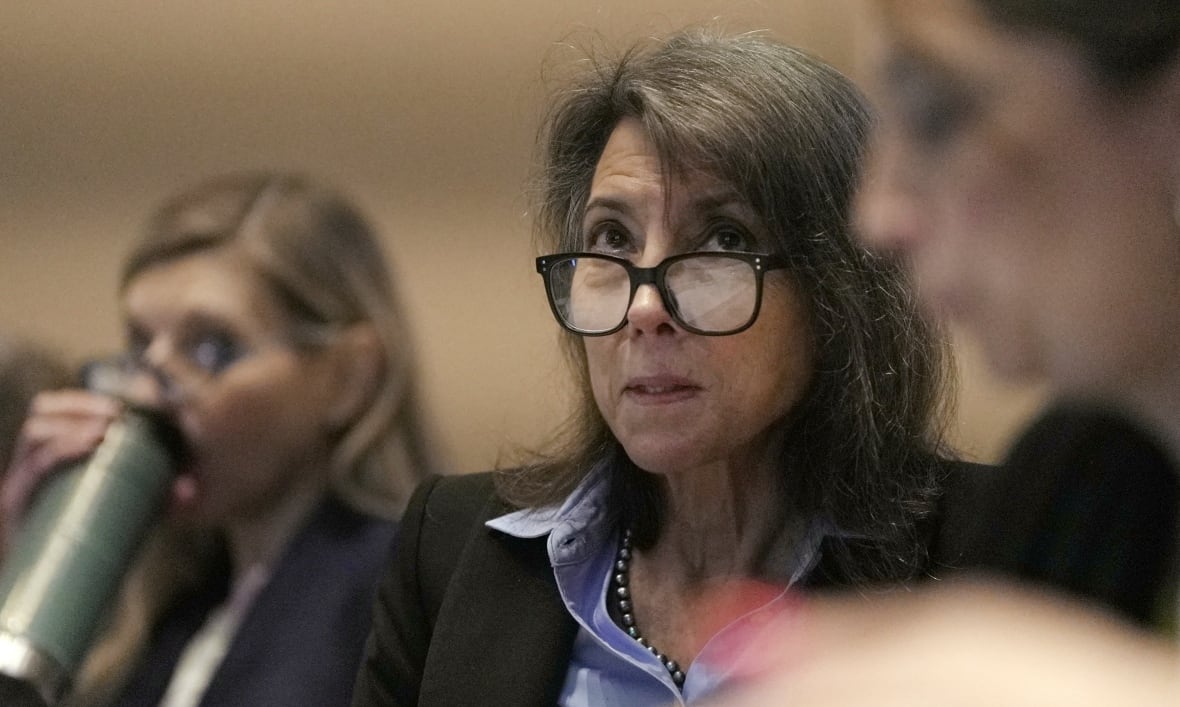 A woman wearing glasses looks up at something above the camera. There are two other people visible in the photo, seemingly sitting near the woman, one drinking from a water bottle.
