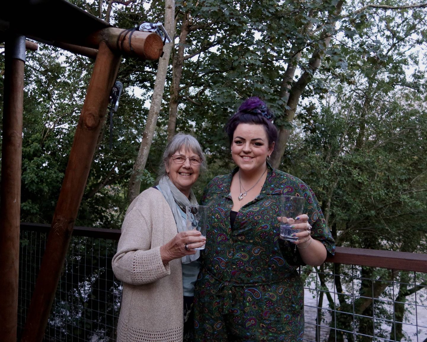 Luisa and her mum stand on the balcony decking of By The Wye amongst the trees next to the River Wye, near Hay-on-Wye. They are enjoying a glass of the gin they made at Silver Circle Distillery earlier that day.
