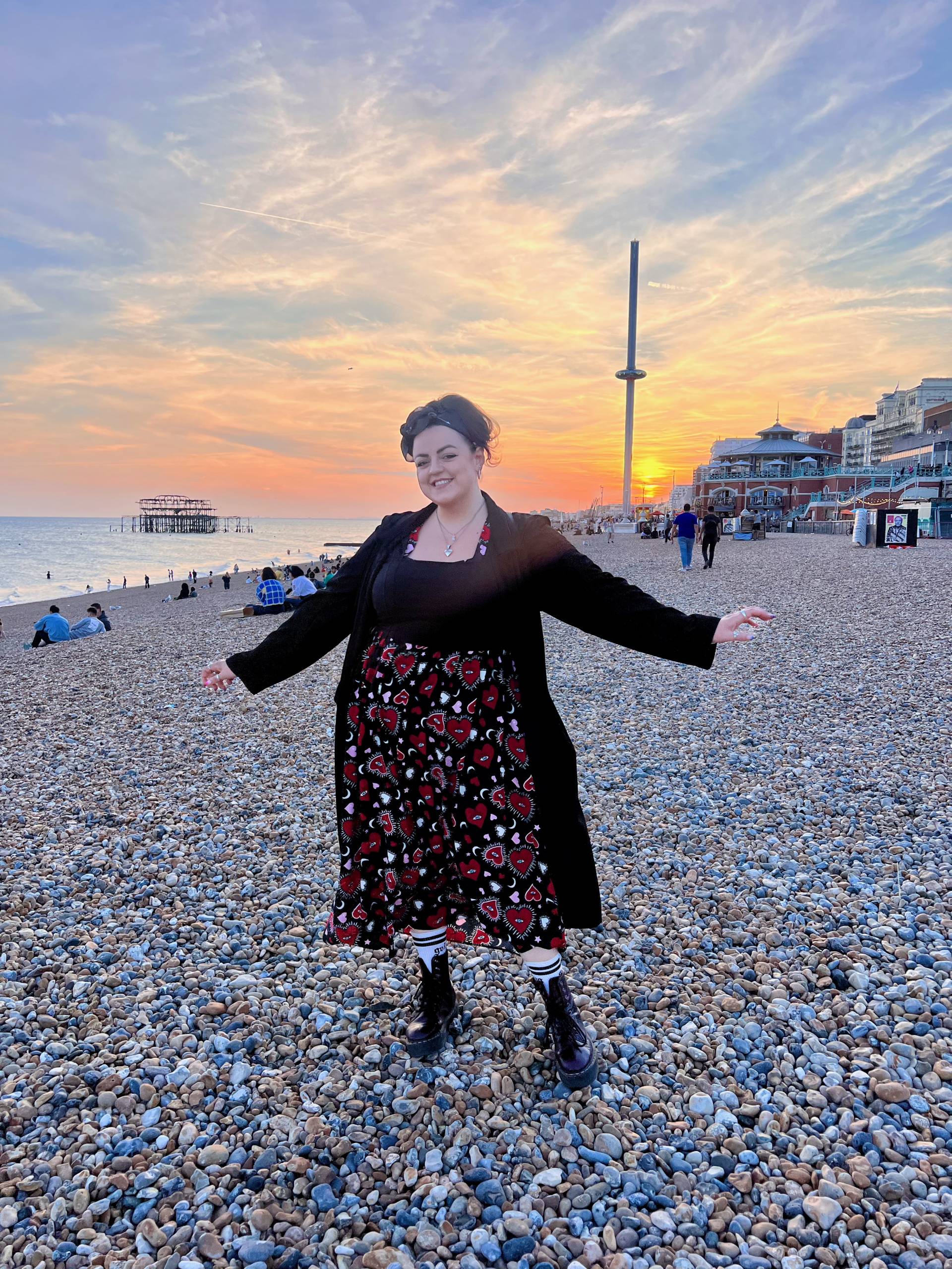 Luisa-Christie stands in front of the i360 on Brighton Beach (which is covered in pebbles). There is a beautiful sunset behind her. She wears a long black and red dress, red Dr Martens and has her arms out, and a big grin on her face.