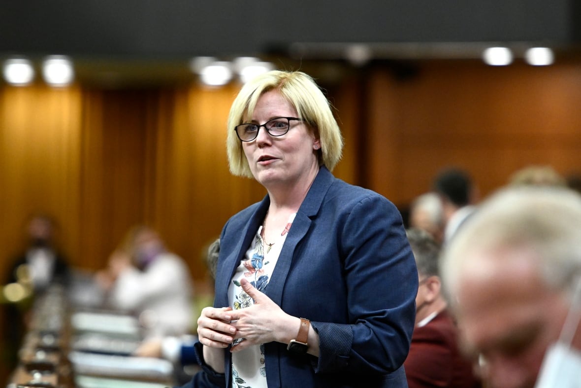 A woman wearing glasses and a blazer stands while speaking in a room filled with people beside and behind her.