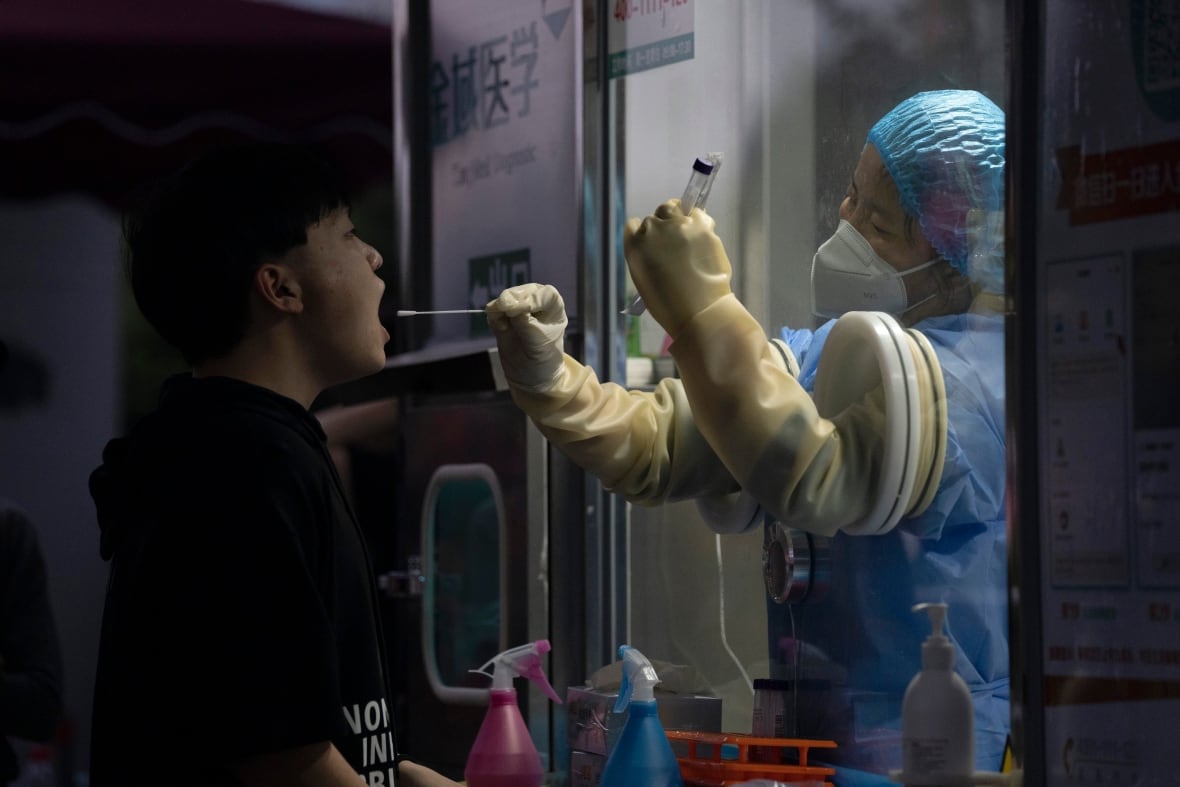 A young man in a black T-shirt has his mouth swabbed by a woman in a medical cap, gown and gloves, standing behind a protective screen.