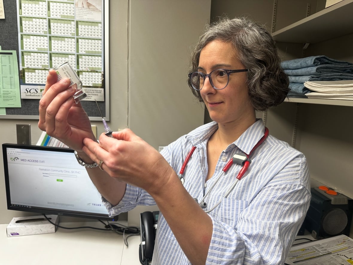 A doctor with a short bob and blue glasses holds a vaccine and a vial. She is standing in a clinic. She is wearing a blue striped button down shirt and wearing a stethoscope around her neck.