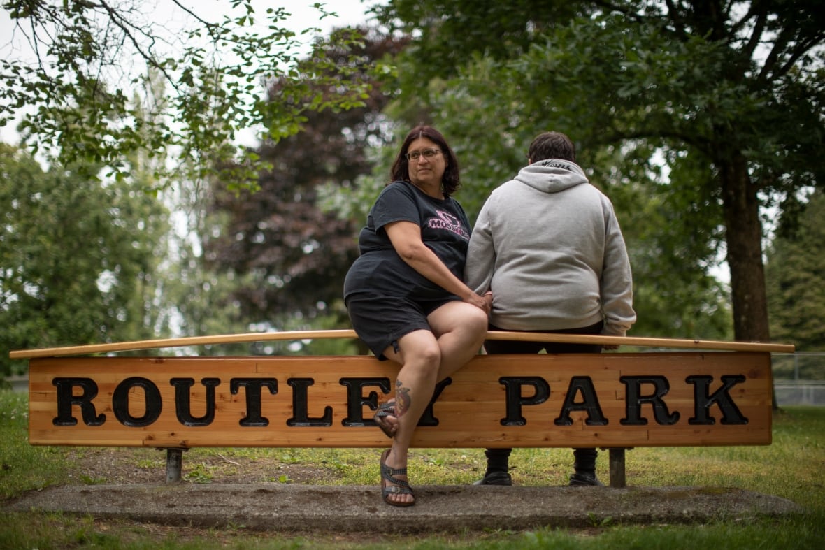 A woman and boy who is turned away sit on a sign that reads 'Routley park.' The woman wears a black T-shirt and black shorts and the boy wears a grey hoodie.