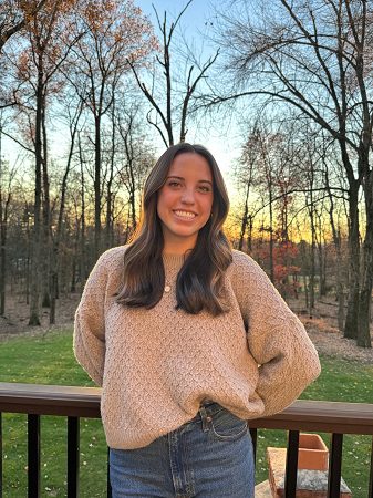 A white woman in pink sweater stands with sunset and trees in background.